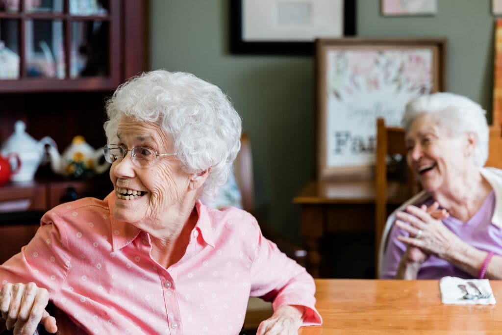 An elderly woman with white hair smiles while sitting at a table indoors.