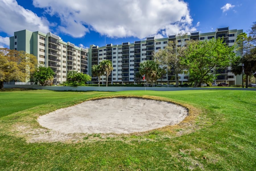 A sand trap on a golf course with a residential building in the background under a partly cloudy sky.