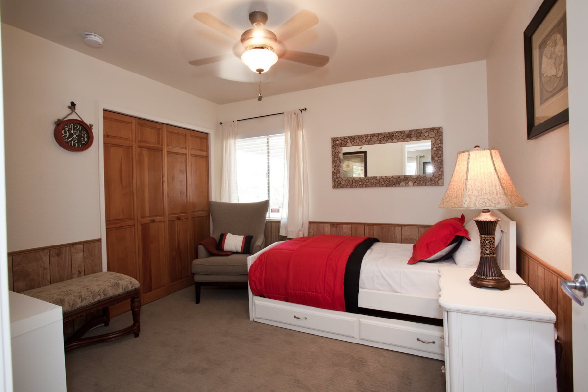 A cozy bedroom featuring a single bed with red bedding, a comfortable armchair, and a lamp on the bedside table. The room has a ceiling fan and closet doors.