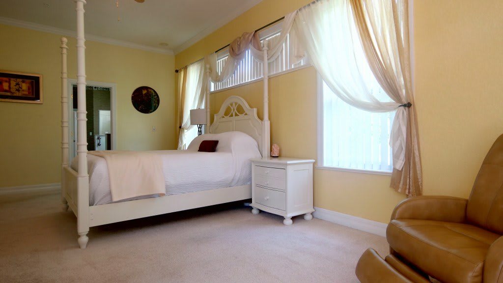 A cozy bedroom interior featuring a white bed and nightstand with light yellow walls and sheer curtains.