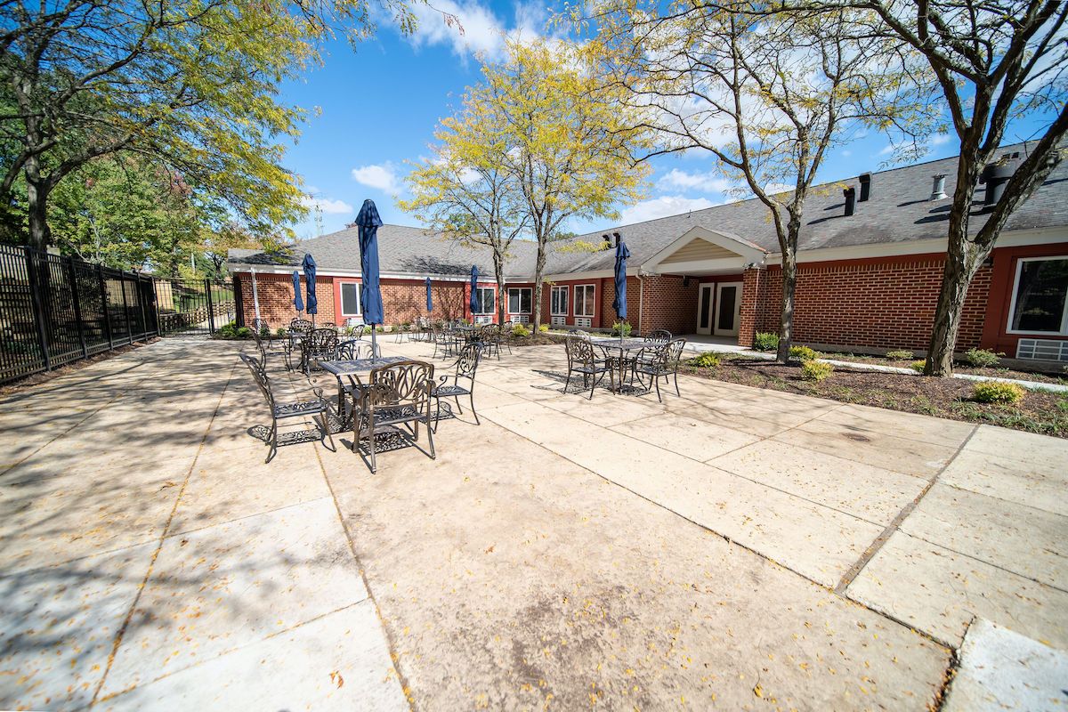 A sunny outdoor patio area with tables and chairs, surrounded by trees and a brick building.
