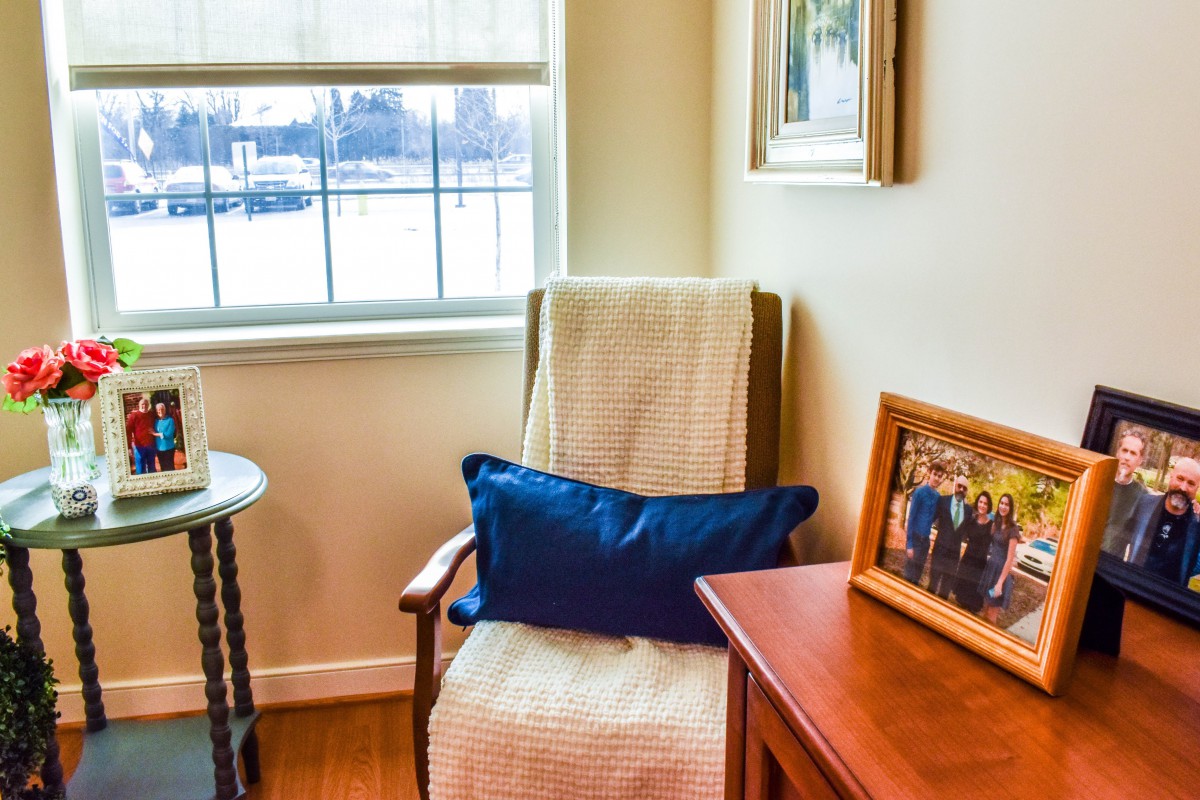 A cozy corner of a living room featuring a chair with a cushion, next to a window and decorated with framed pictures.