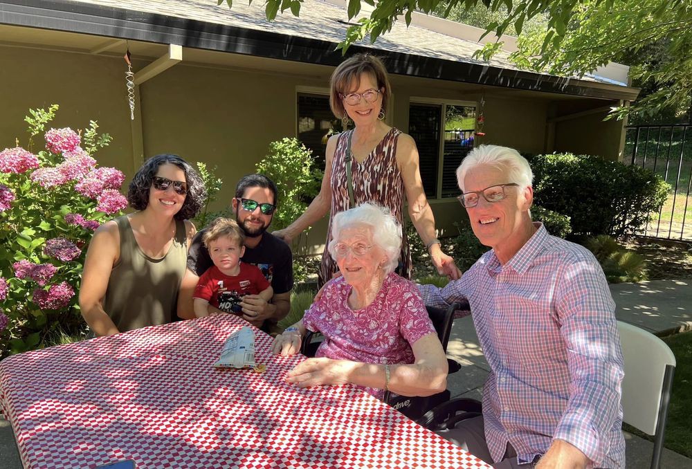A multi-generational family gathering outdoors, seated around a checkered tablecloth.
