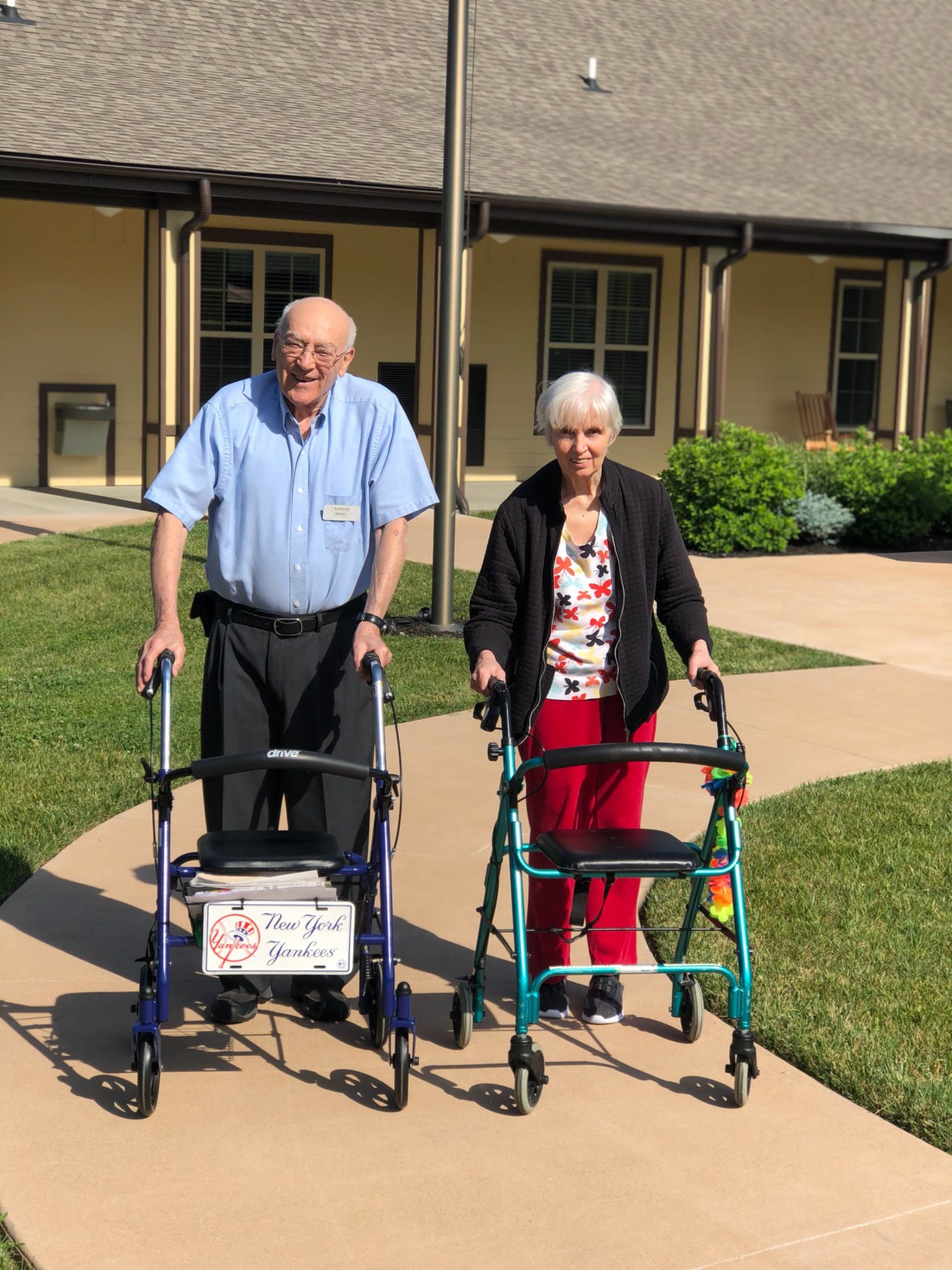 An elderly couple using walkers outside a building on a sunny day.