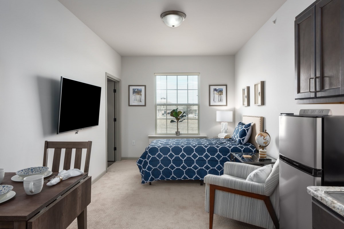 A modern bedroom with blue patterned bedding, a wall-mounted TV, and ample natural light from the window.