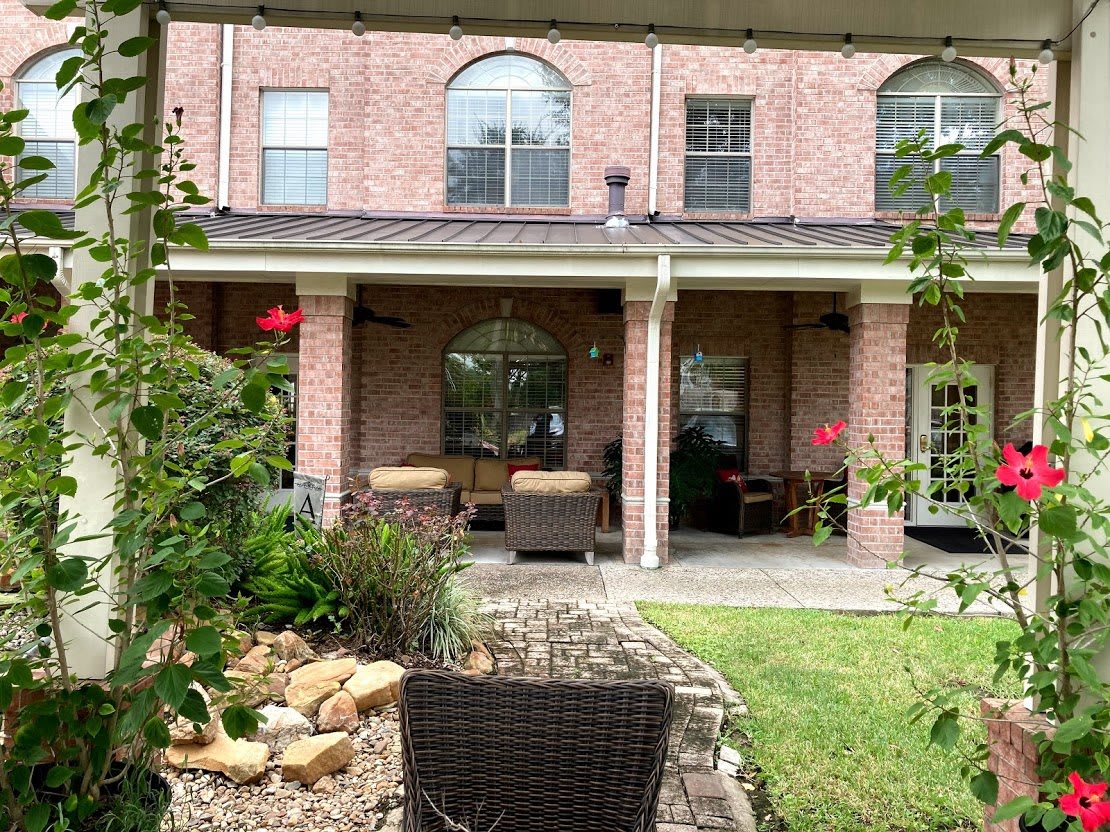 A peaceful backyard patio with comfortable outdoor seating and well-maintained garden in front of a brick house.