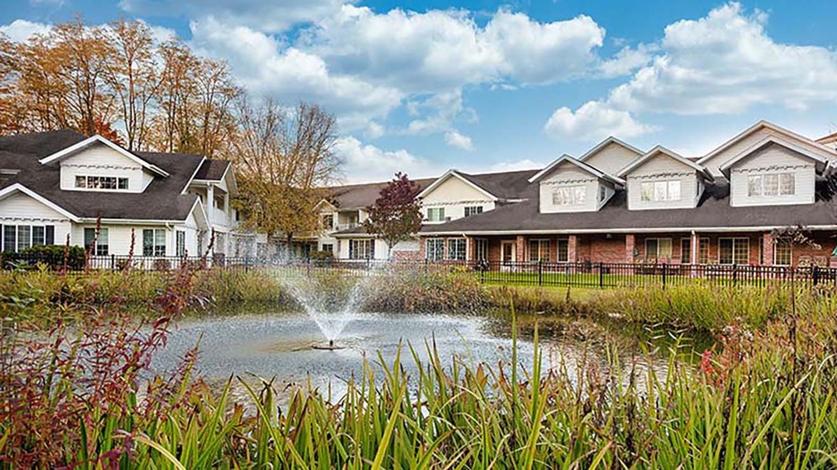 A serene view of houses by a pond with a fountain under a partly cloudy sky.