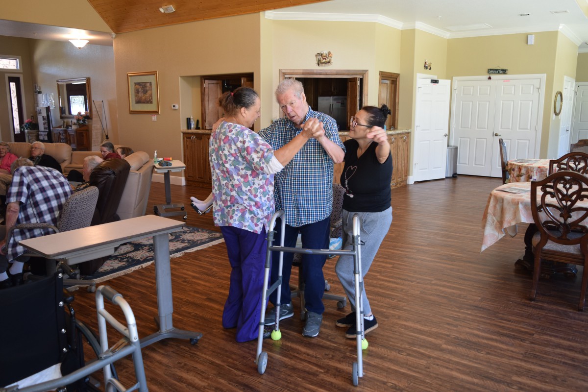 Caregivers assist an elderly man with a walker, providing support and encouragement.