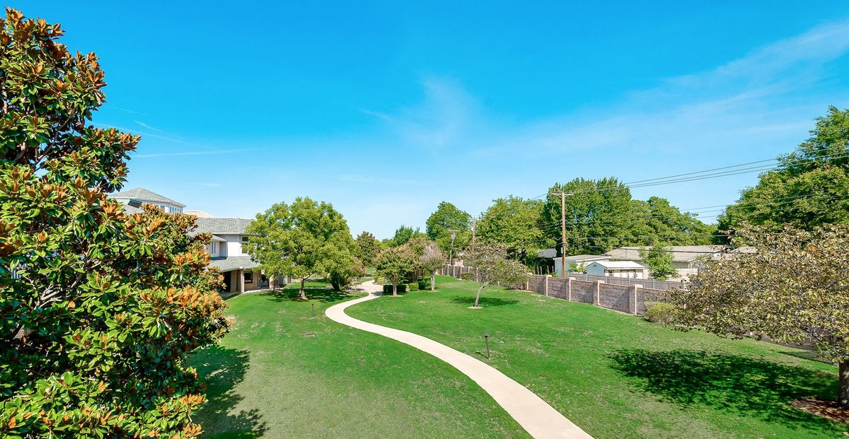 A scenic view of a park featuring a winding pathway surrounded by lush green grass and trees under a clear blue sky.