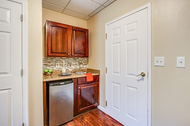 A small kitchenette with wooden cabinets, a compact refrigerator, and a sink set against a decorative backsplash.
