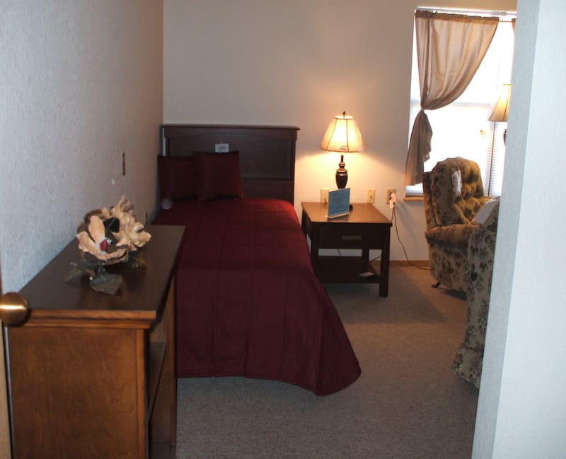A cozy bedroom with a single bed, nightstand, and a comfortable chair near the window.