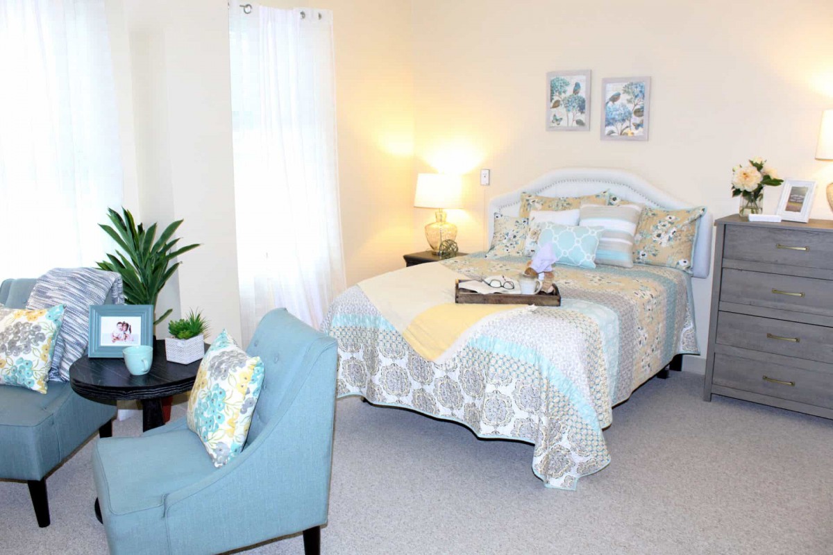 A cozy and well-decorated bedroom featuring a neatly made bed with decorative pillows, a small bedside lamp, an armchair, and soft lighting.