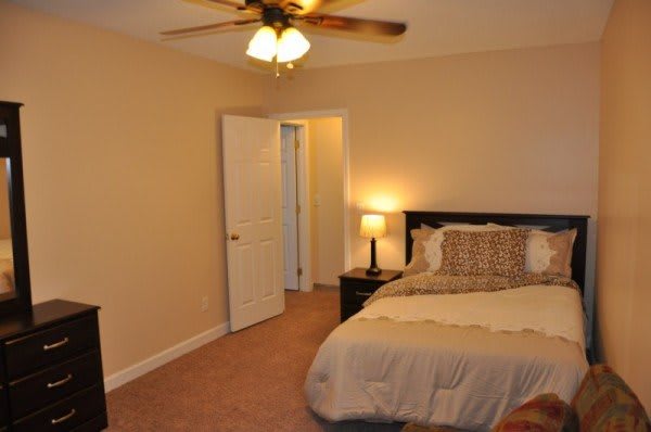 A cozy bedroom featuring a neatly made bed, a nightstand with a lamp, beige walls, and a ceiling fan.