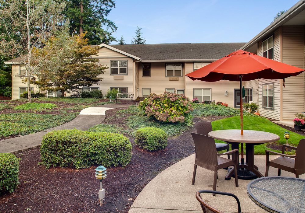 A peaceful garden patio with seating and an umbrella, surrounded by manicured landscaping and adjacent to a residential building.