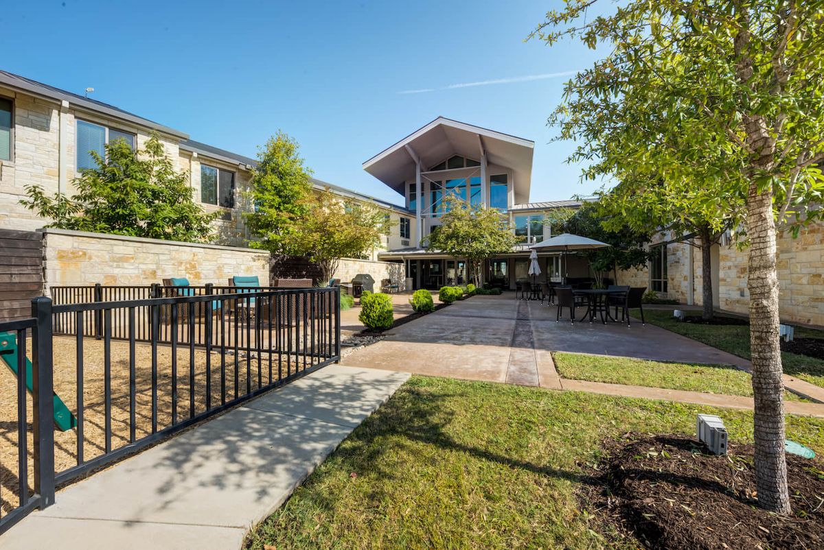 A modern building with a garden and pathway, surrounded by trees under a clear blue sky.