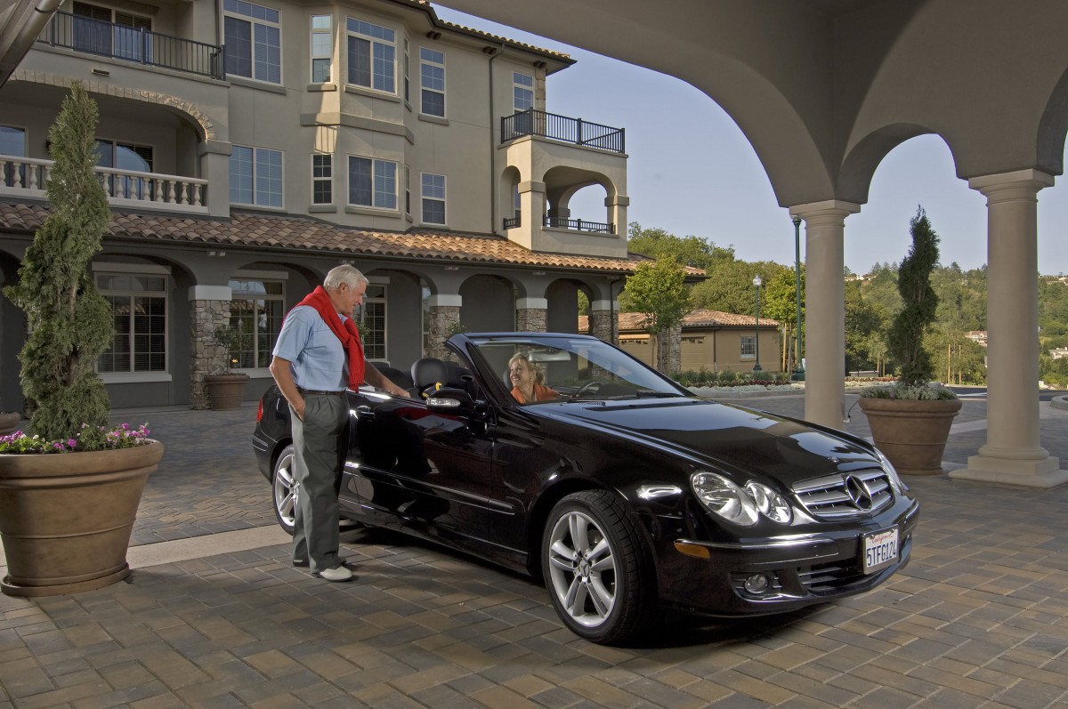 A man stands beside a black Mercedes-Benz in front of a luxurious house.