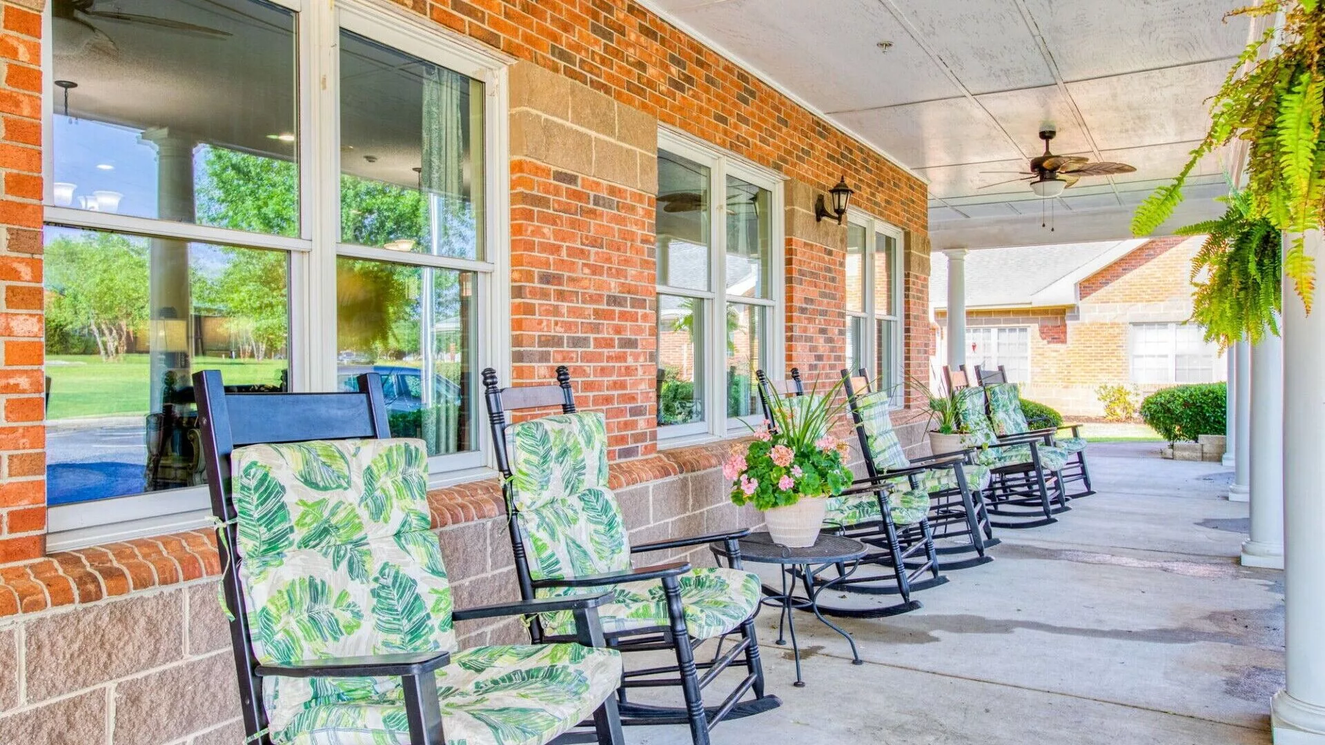 A row of rocking chairs with patterned cushions on a porch of a brick building, accompanied by potted plants.
