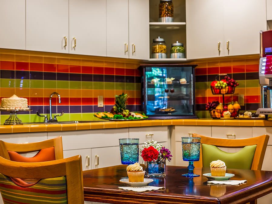A vibrant kitchen features colorful striped tiles, a cozy dining area with cupcakes and glassware, accented by fresh flowers and decorative elements.