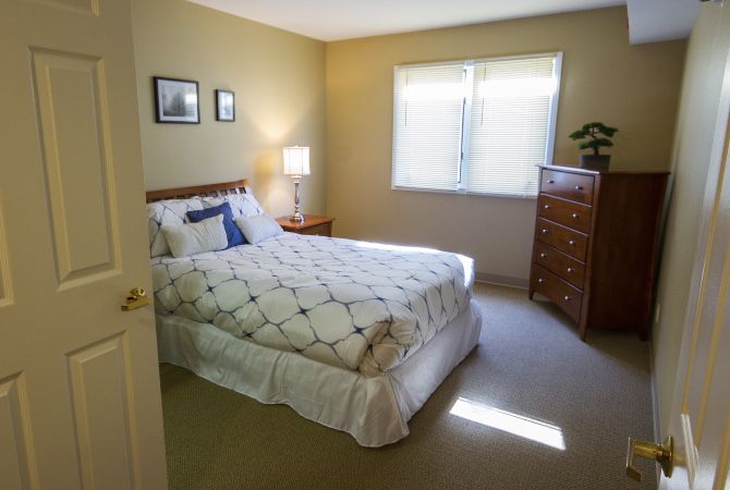 A cozy bedroom with a neatly made bed, side table with a lamp, and a window bringing in natural light.