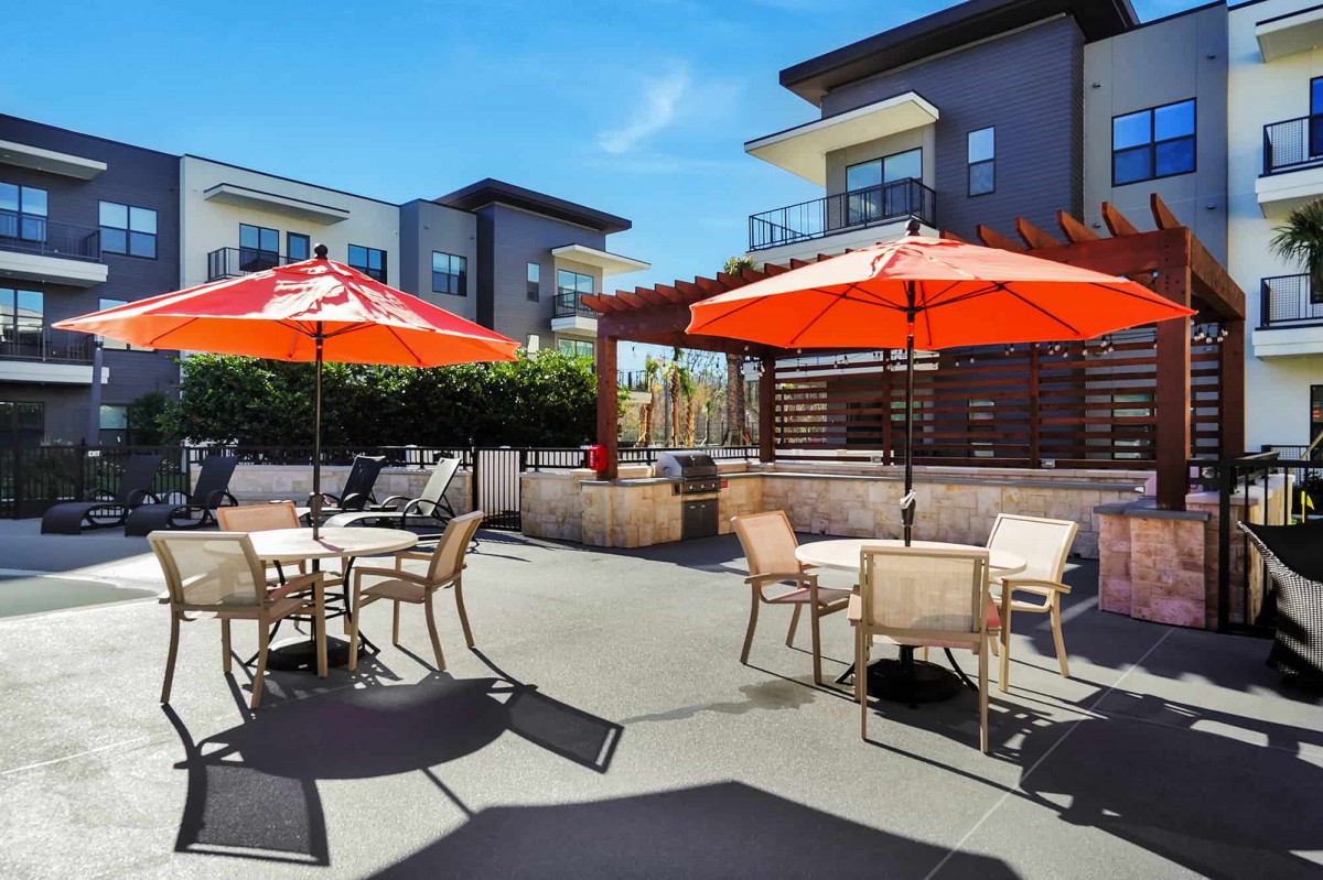 A modern outdoor patio area with tables, chairs, and orange umbrellas under a clear blue sky.
