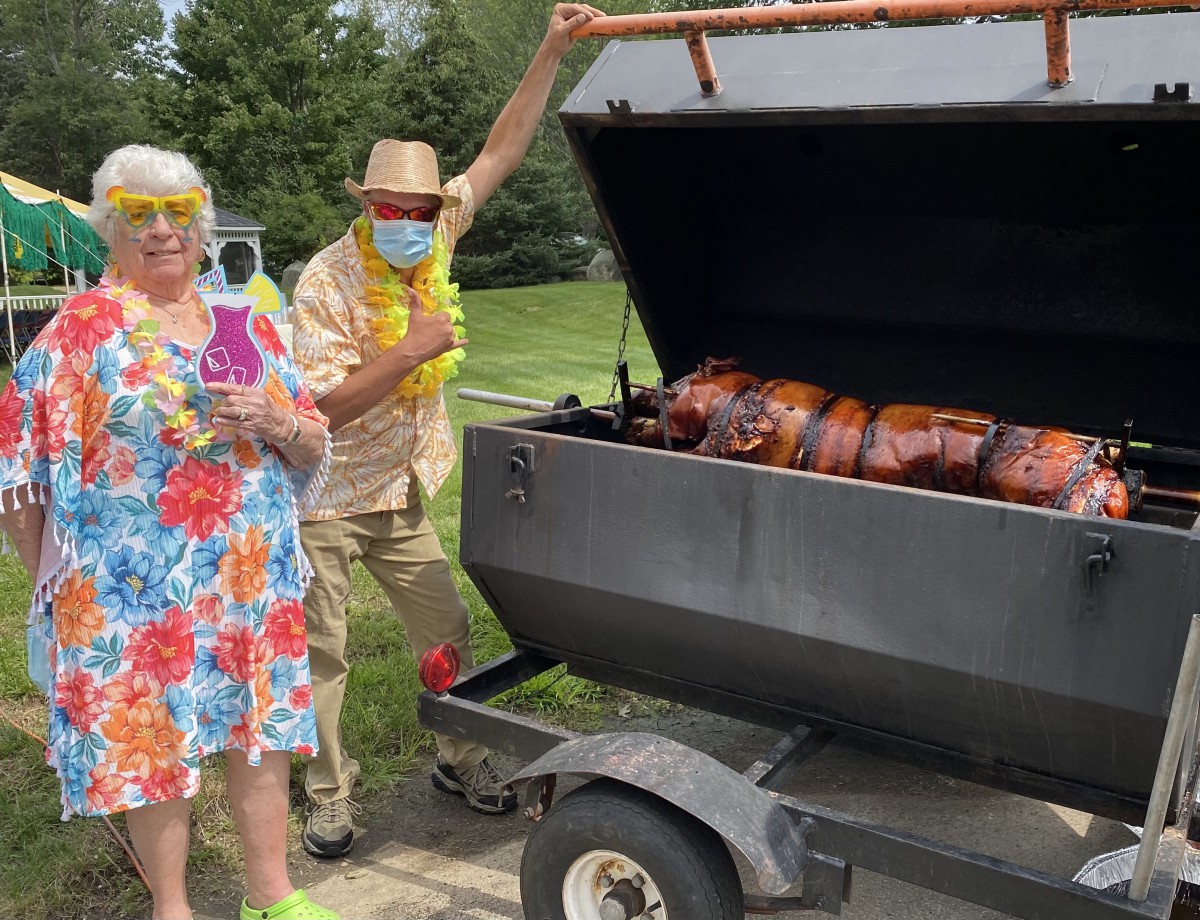 Two people dressed in festive clothing stand by a large outdoor grill with roasted meat.
