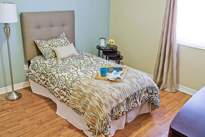 A cozy bedroom with a neatly made bed, decorated with patterned bedding and pillows. A tray with items is placed on top, and a lamp sits on the bedside table.