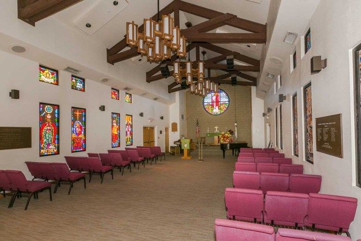 Interior view of a chapel with stained glass windows, wooden beams, and pink pews.