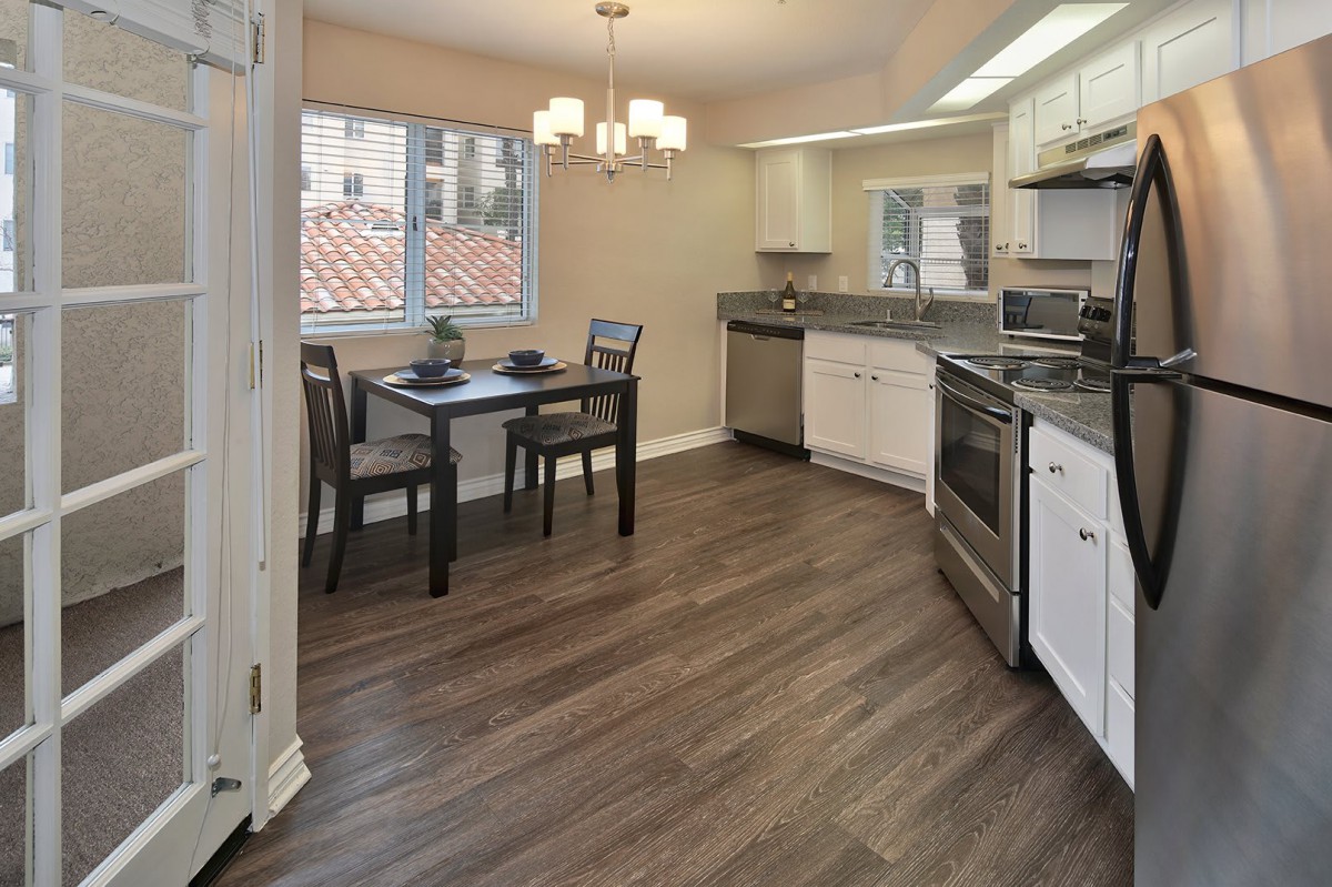A modern kitchen with hardwood floors, stainless steel appliances, and a small dining area by a large window.