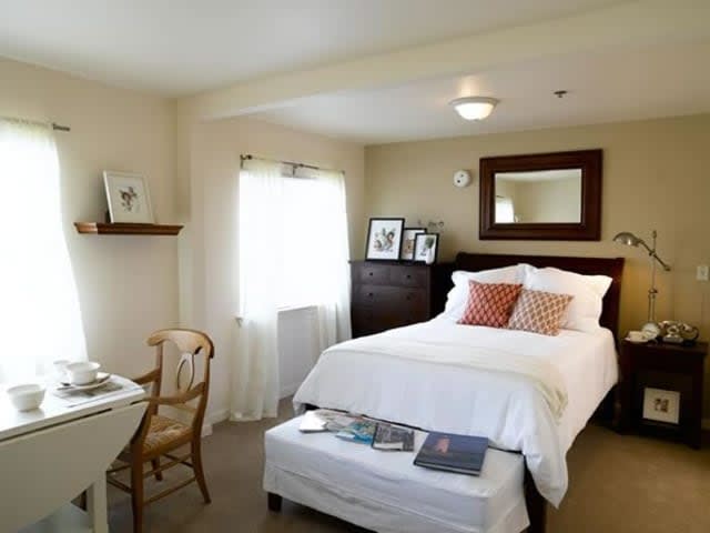 A cozy bedroom with a neatly made bed, mirror, and natural light from the window.