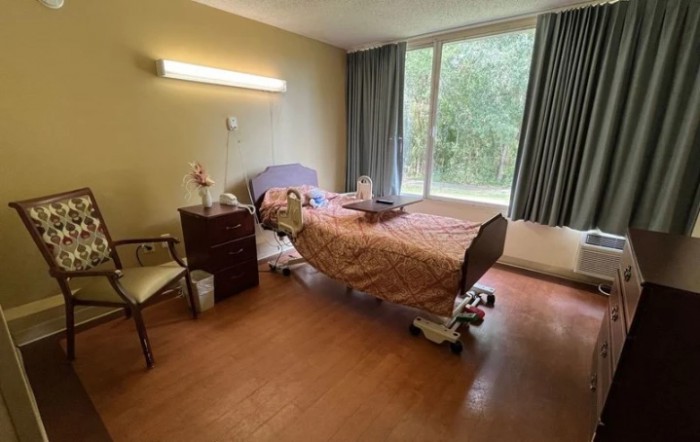 A hospital room with a single bed, nightstand, and a chair by the window.
