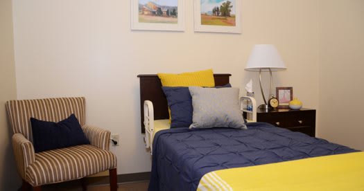 A cozy bedroom with a neatly made bed featuring blue and yellow bedding, accompanied by a bedside lamp and a striped armchair. Two framed artworks hang on the wall above the headboard.