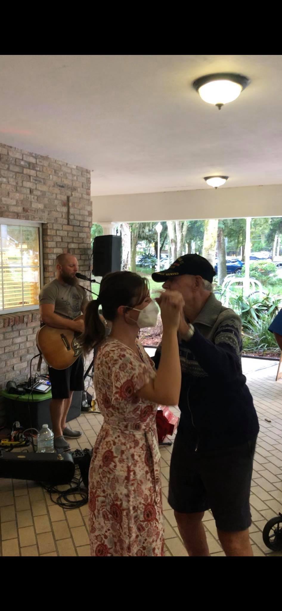 An elderly man dances with a woman in a floral dress while live music plays in the background.