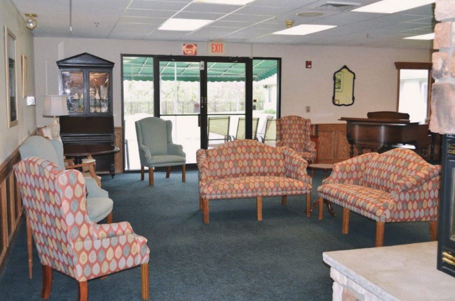 A lounge area with various chairs and sofas arranged on a carpeted floor, featuring large windows in the background.