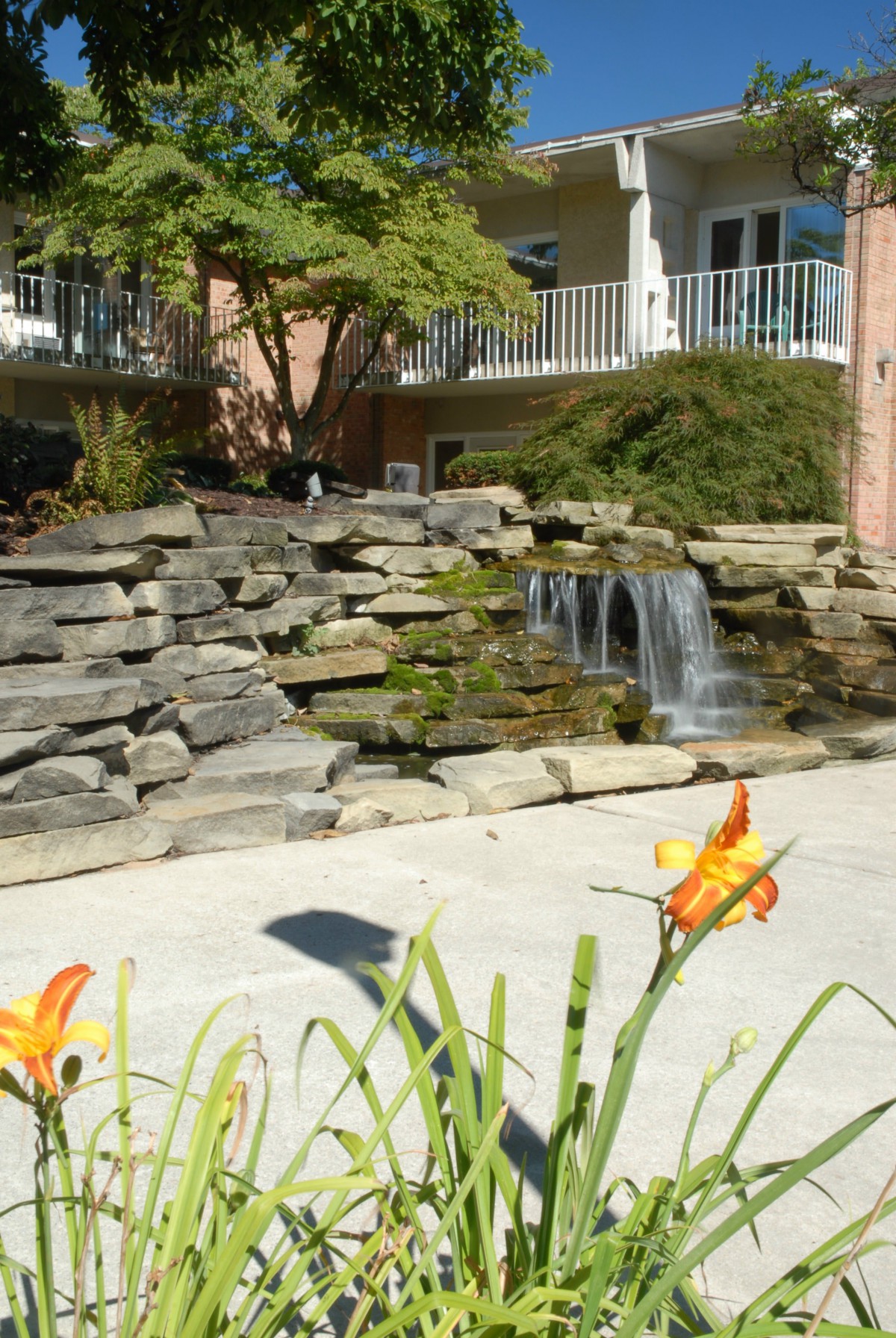 A small waterfall cascades over rocks in a landscaped garden, adorned with orange flowers.