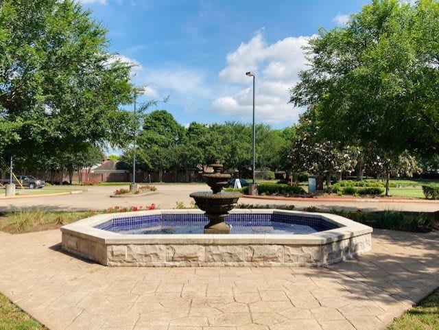 A stone fountain in a landscaped outdoor setting with trees and a clear blue sky.