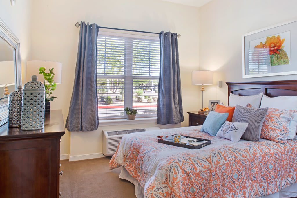 A cozy bedroom with a neatly made bed, large window with gray curtains, and a bedside lamp.