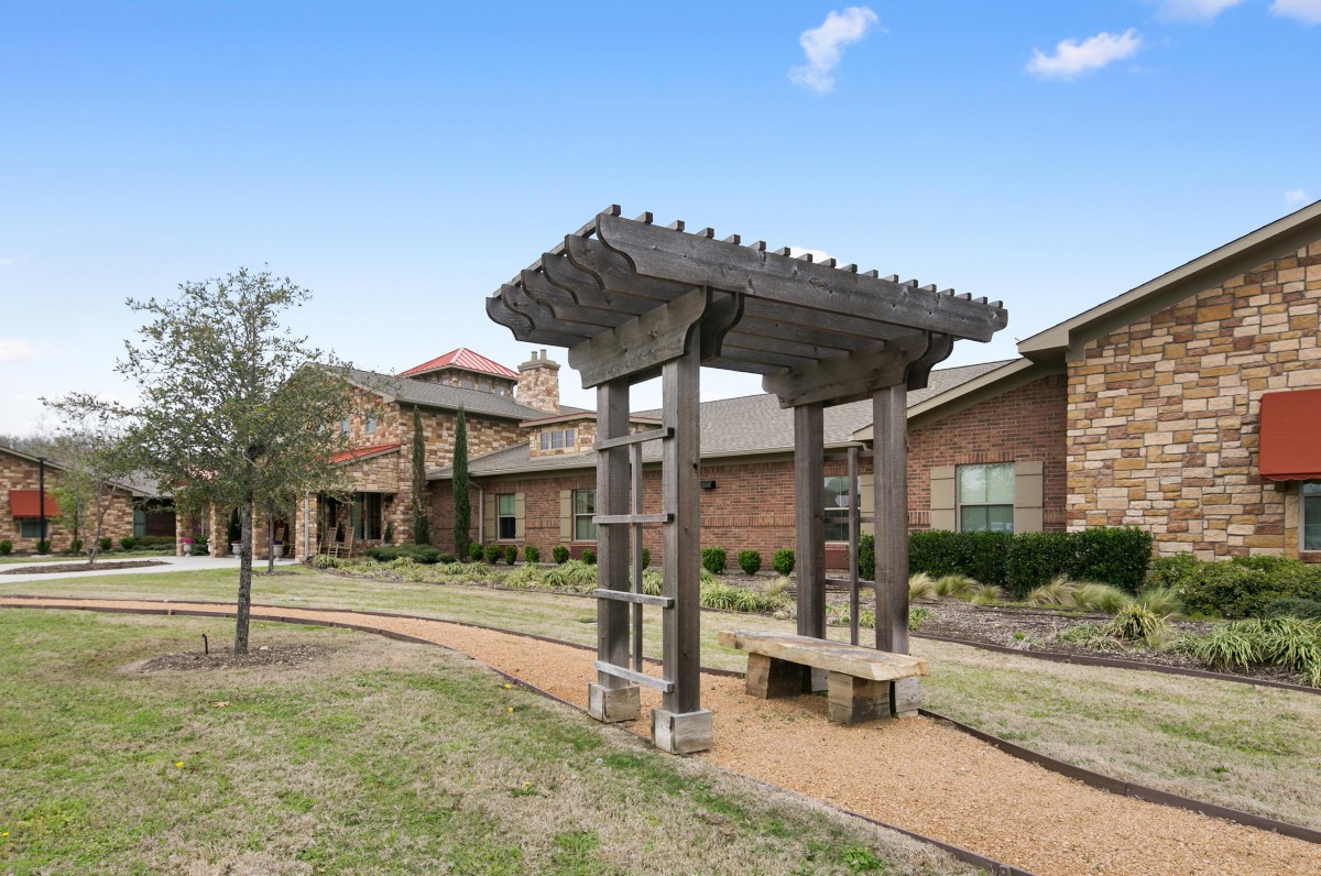 A garden area featuring a wooden pergola along a gravel pathway with a building in the background.