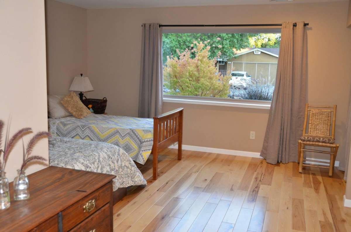 A cozy bedroom with a large window, wooden floor, and simple furnishings.