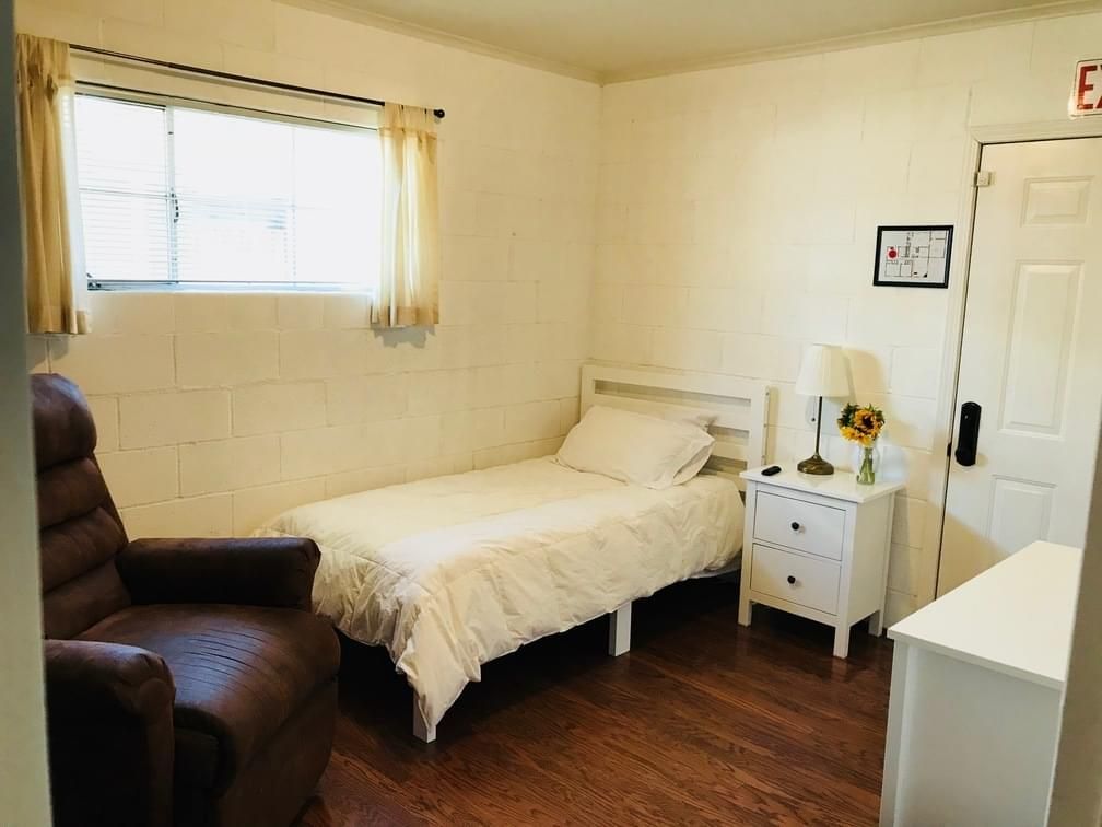 A simple and cozy bedroom with a single bed, nightstand, lamp, and a window letting in natural light.