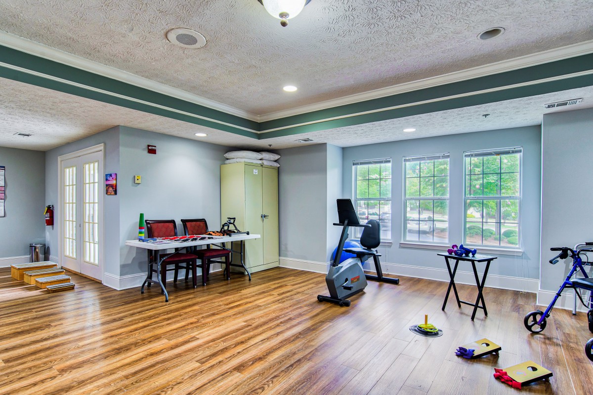 A spacious exercise room with a stationary bike, desk, and chairs on wood flooring.
