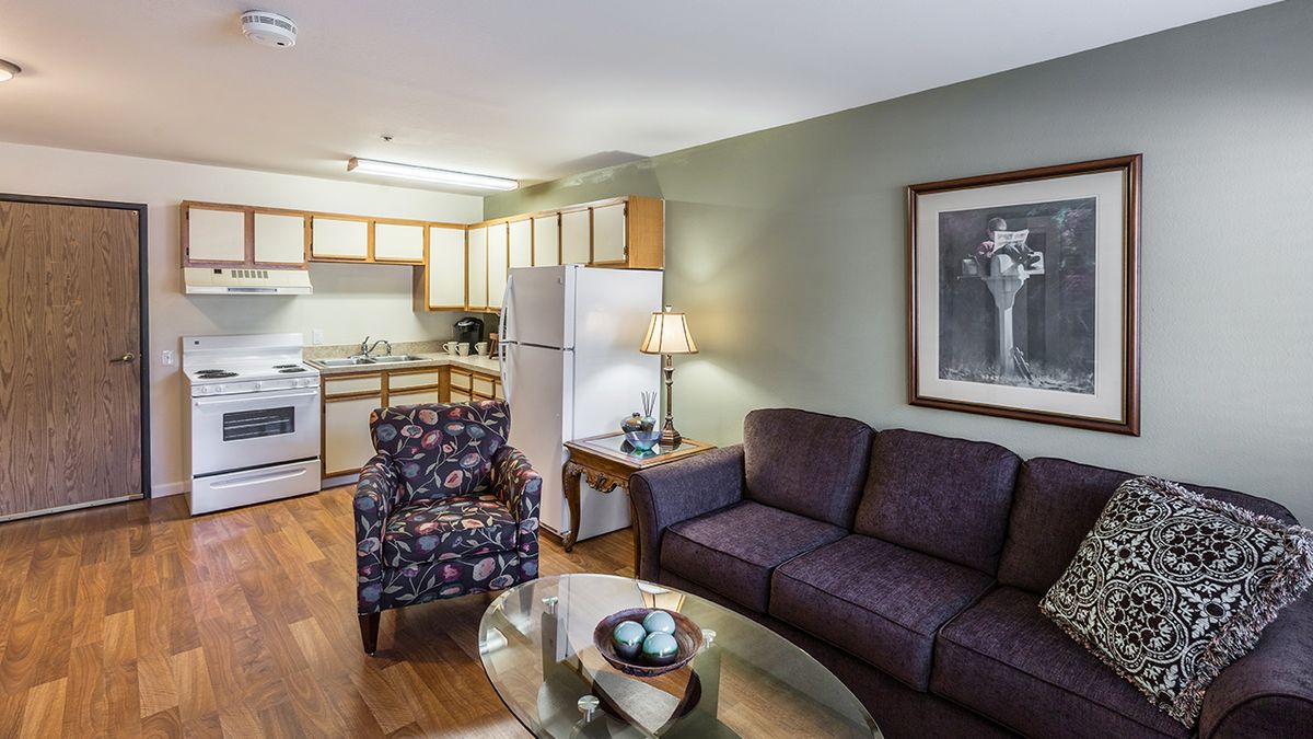 A cozy living room with an adjacent kitchen, featuring a purple couch, patterned armchair, and modern appliances.