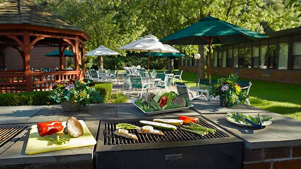 An outdoor barbecue setup with vegetables on the grill, surrounded by tables and umbrellas in a garden setting.