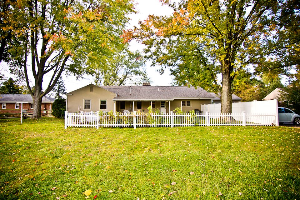 A single-story house with a white fence and a spacious yard surrounded by trees in autumn foliage.