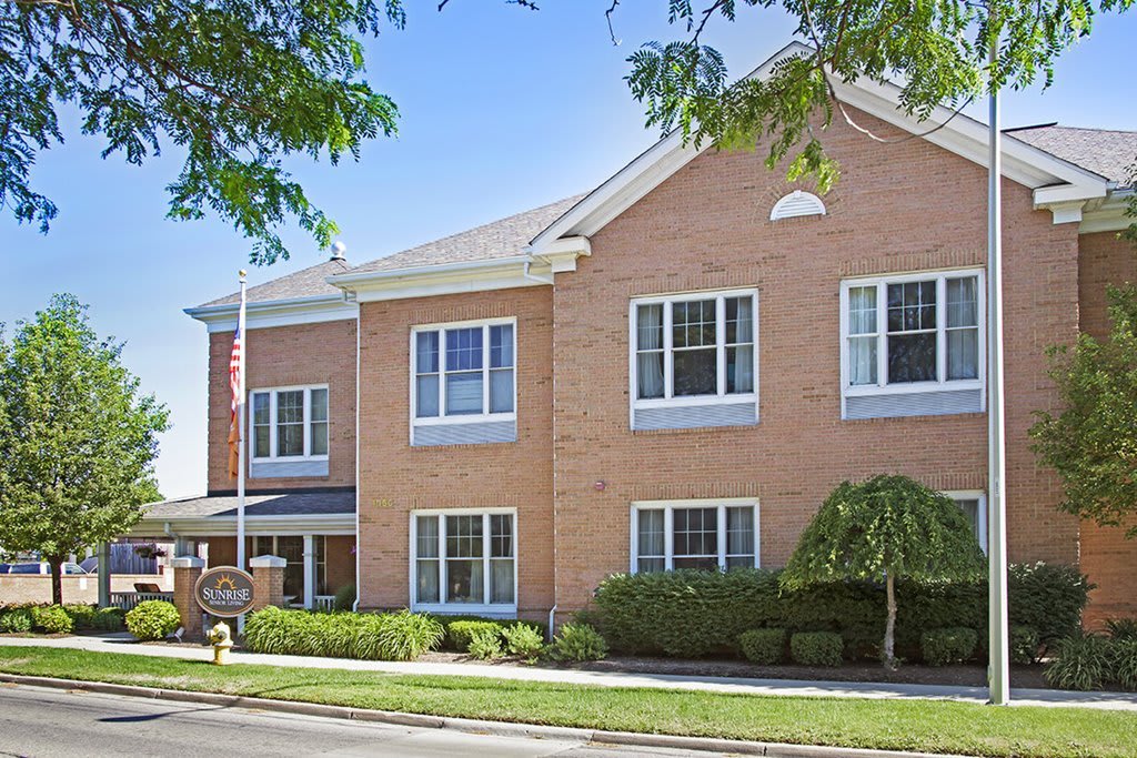 A two-story brick building with windows, surrounded by trees and landscaped greenery, featuring a flagpole at the entrance.
