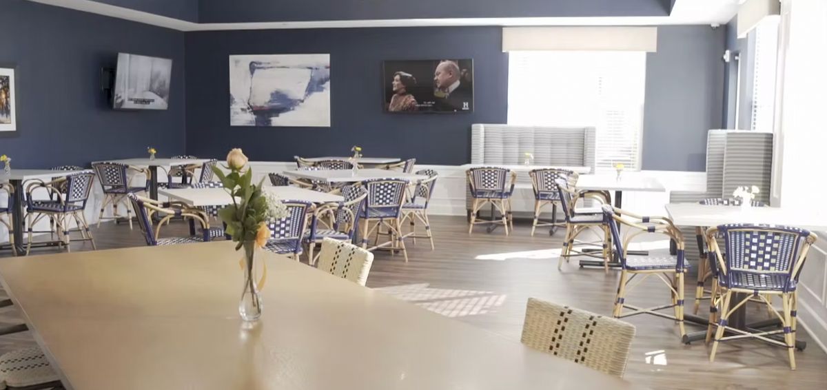 A cozy cafe interior with empty tables and chairs, decorated with flowers and artwork on the walls.
