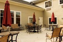 An inviting patio area with tables and chairs, shaded by red umbrellas, against a light-colored building exterior.