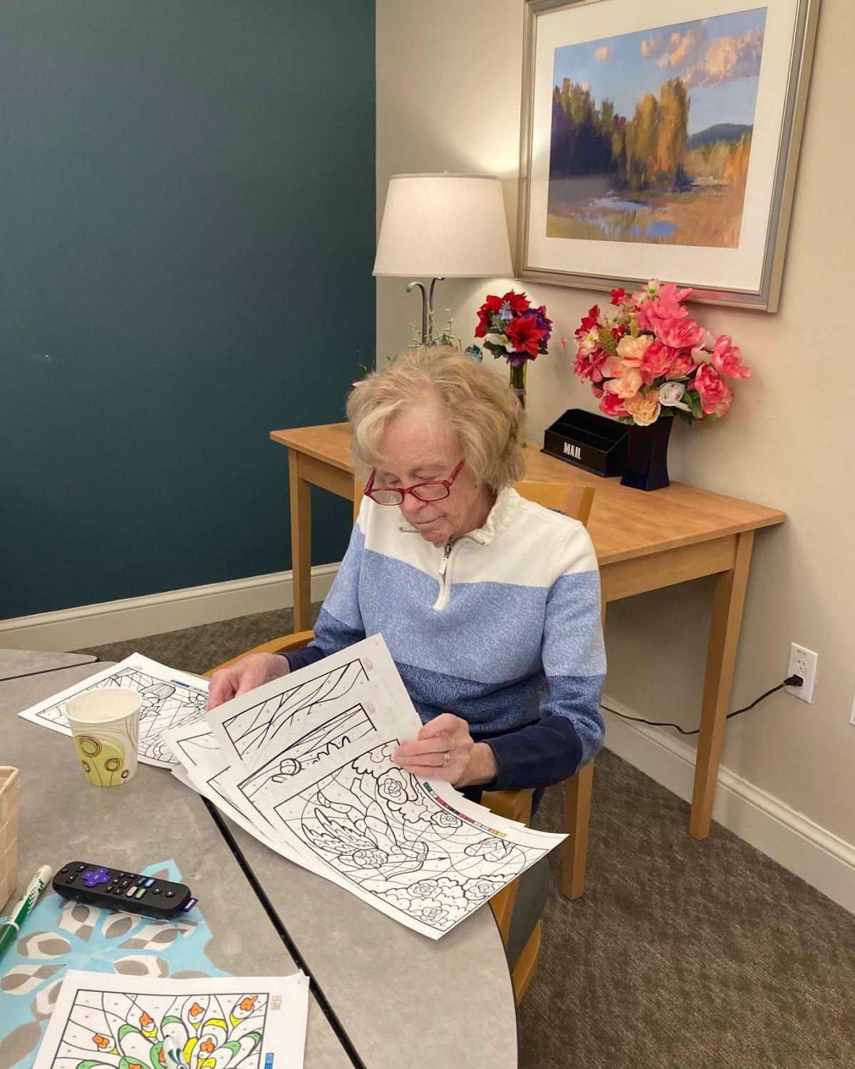 A woman is sitting at a table indoors, reading a book with flowers and a lamp in the background.