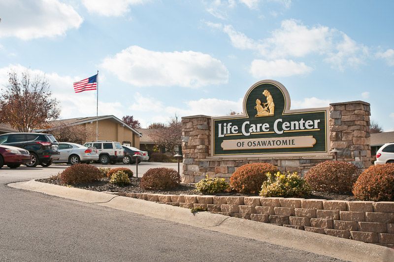 A nursing home facility with a sign, parking lot, and American flag in the background.