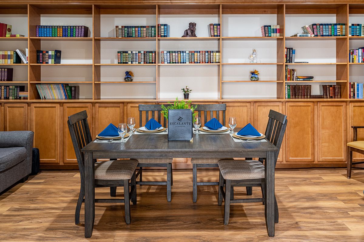 A well-set dining table with blue napkins, surrounded by chairs, in front of a wooden bookshelf filled with books.