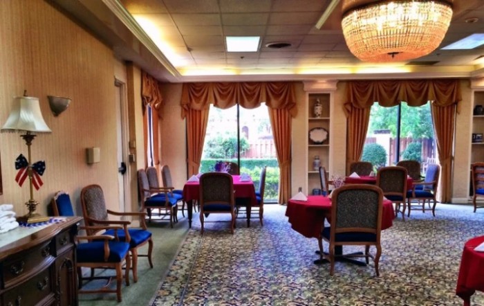 A formal dining room with elegantly set tables and chairs, featuring a chandelier and large windows with draped curtains.